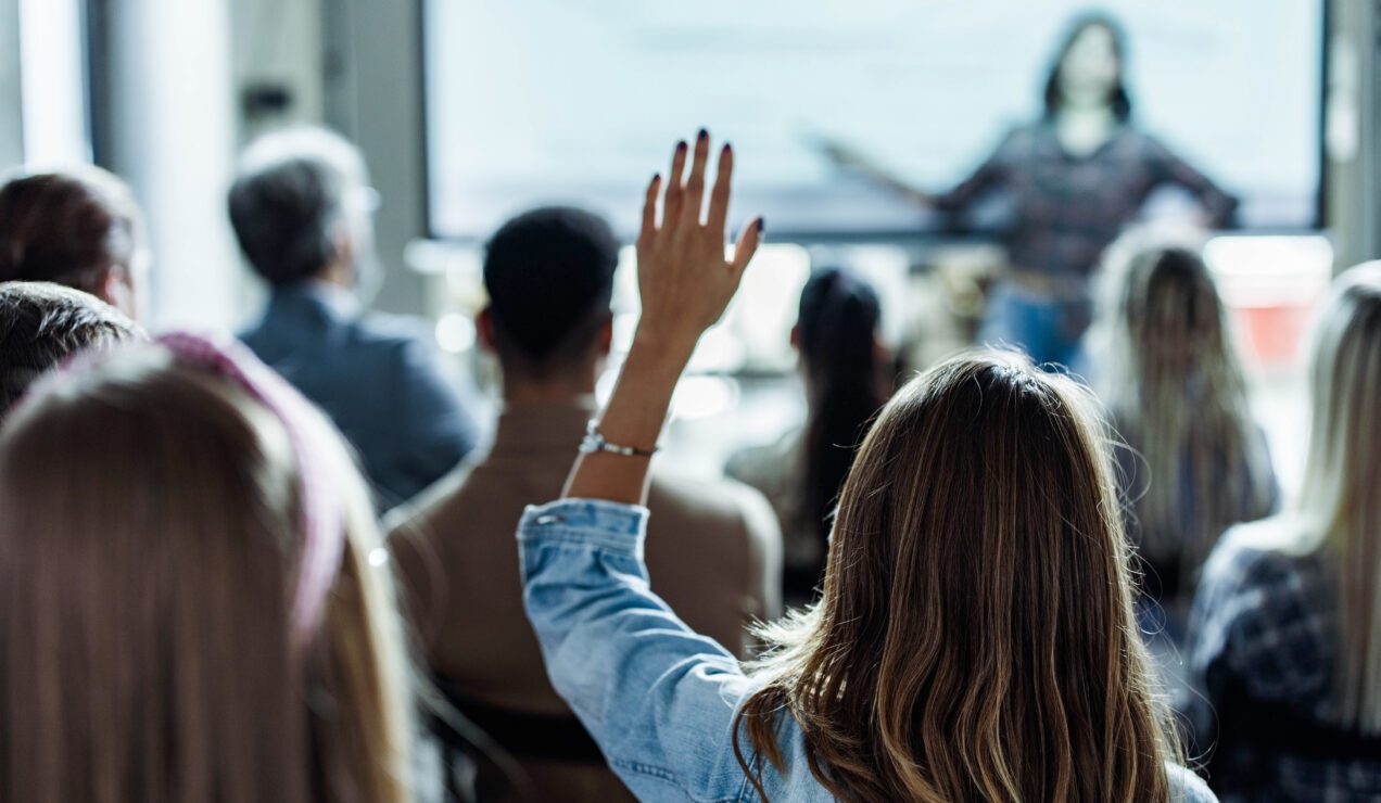 Rear view of a female freelancer raising her hands to ask a question on a seminar in board room.